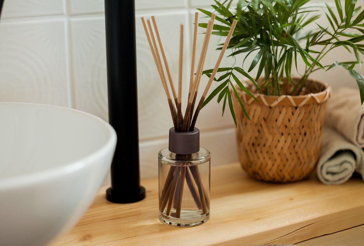 A reed diffuser a potted plant a rolled towel and a sink faucet on a wooden bathroom counter