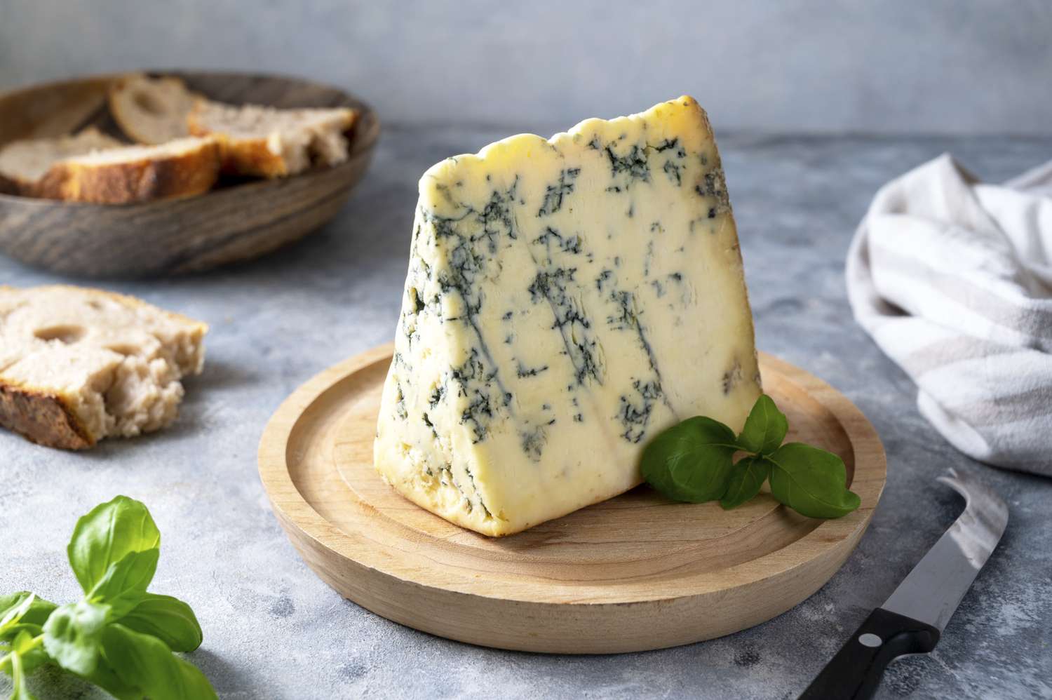 A wedge of blue cheese with green leaves on a wooden platter bread in a bowl and a knife are nearby on the table