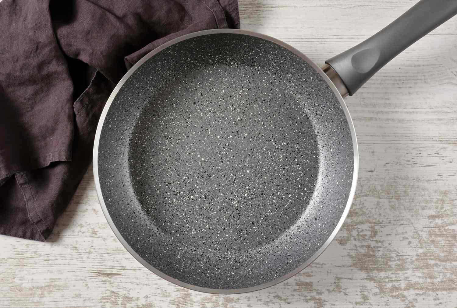 A nonstick frying pan with a speckled surface placed on a wooden surface next to a fabric cloth