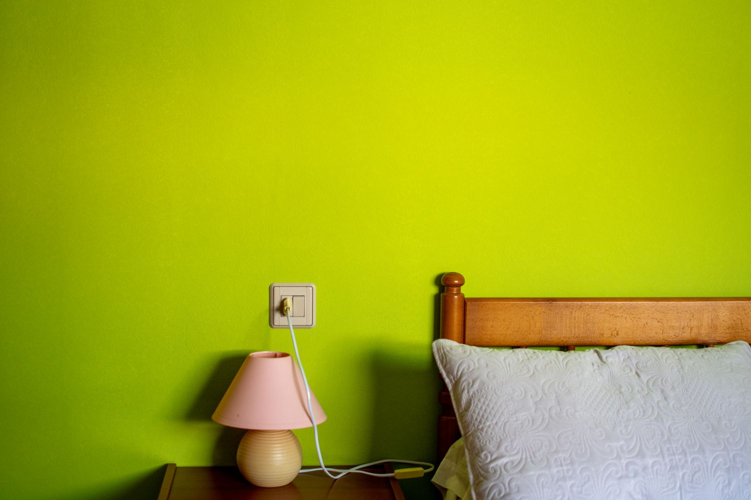 A wooden bed frame with a pillow next to a nightstand holding a pink lamp and an electrical outlet on a lime green wall
