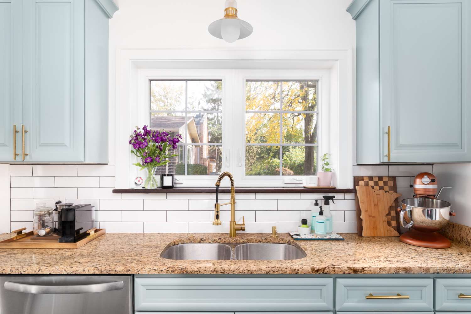 A modern kitchen sink setup with a double bowl and a view out of a window featuring cabinetry and kitchen appliances