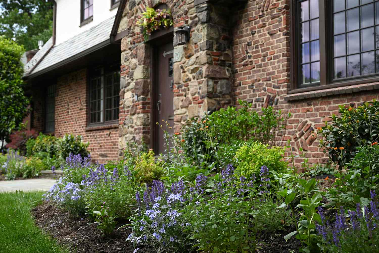A flower garden with blooming plants next to a brick house with a stone and wood exterior