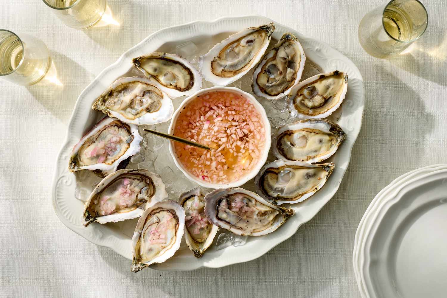 A platter of oysters on ice with a bowl of mignonette sauce in the center surrounded by glasses of a drink on a white tablecloth