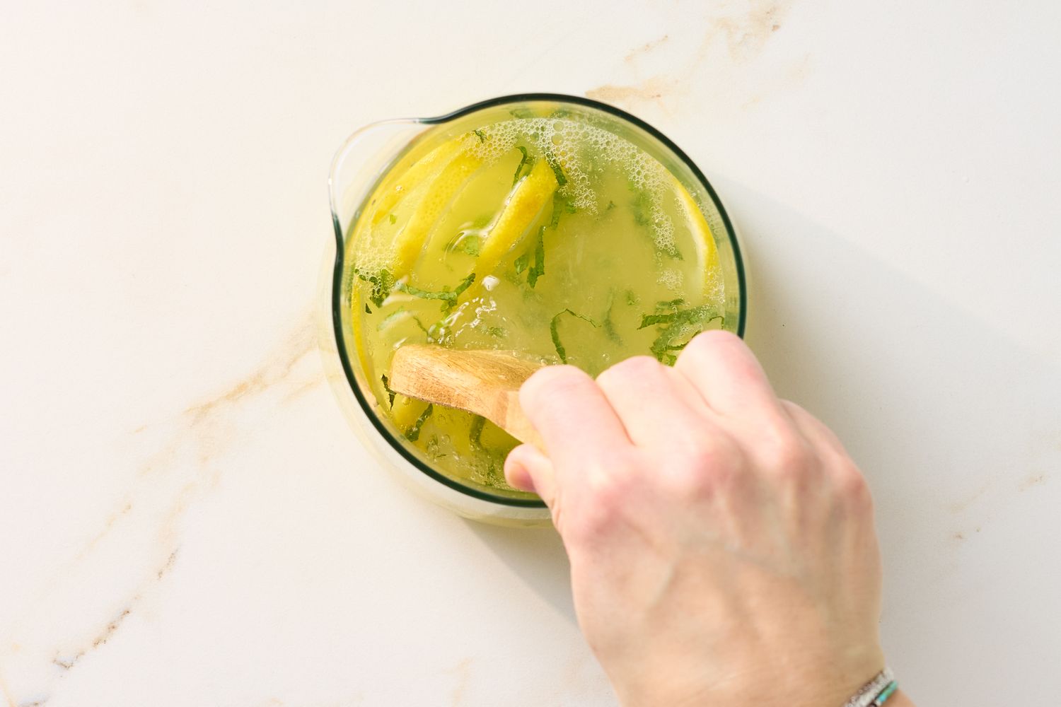 A hand stirring mint lemonade in a glass pitcher with a wooden spoon