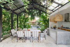 Outdoor space with a dining table chairs a kitchen counter and vegetation around under a glassroofed pergola