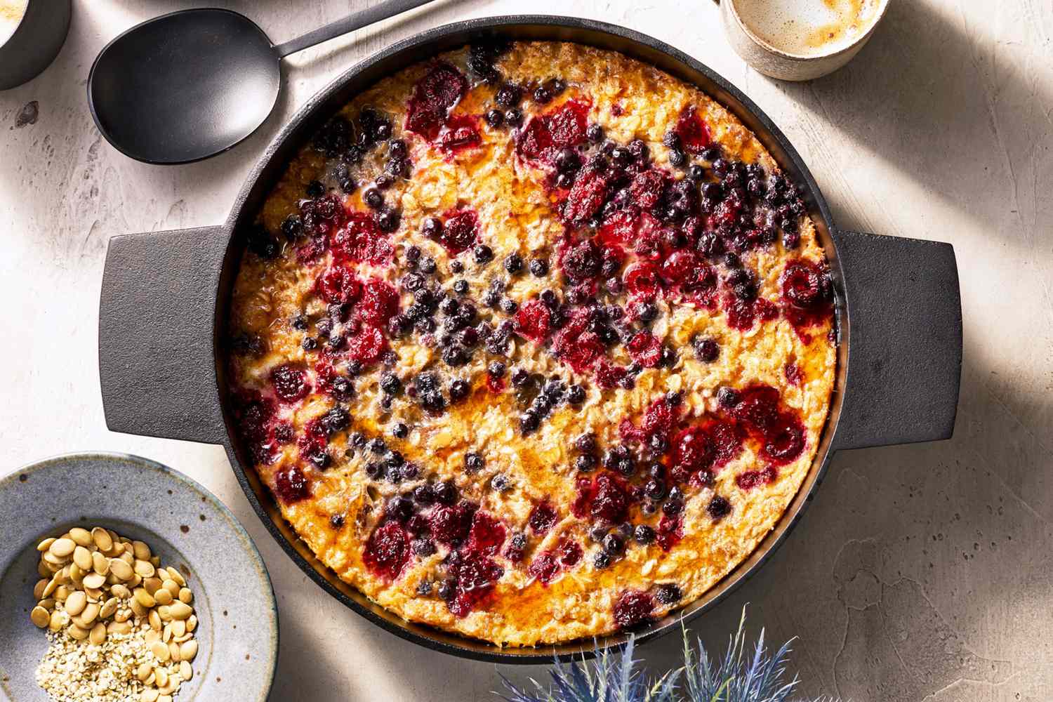 A baked oatmeal dessert garnished with berries in a round baking dish surrounded by serving utensils and small serving items on a table