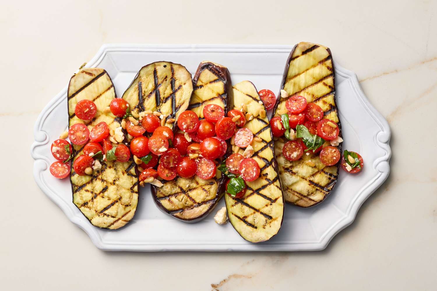 Grilled eggplant slices topped with cherry tomatoes, basil, and feta cheese, served on a rectangular platter