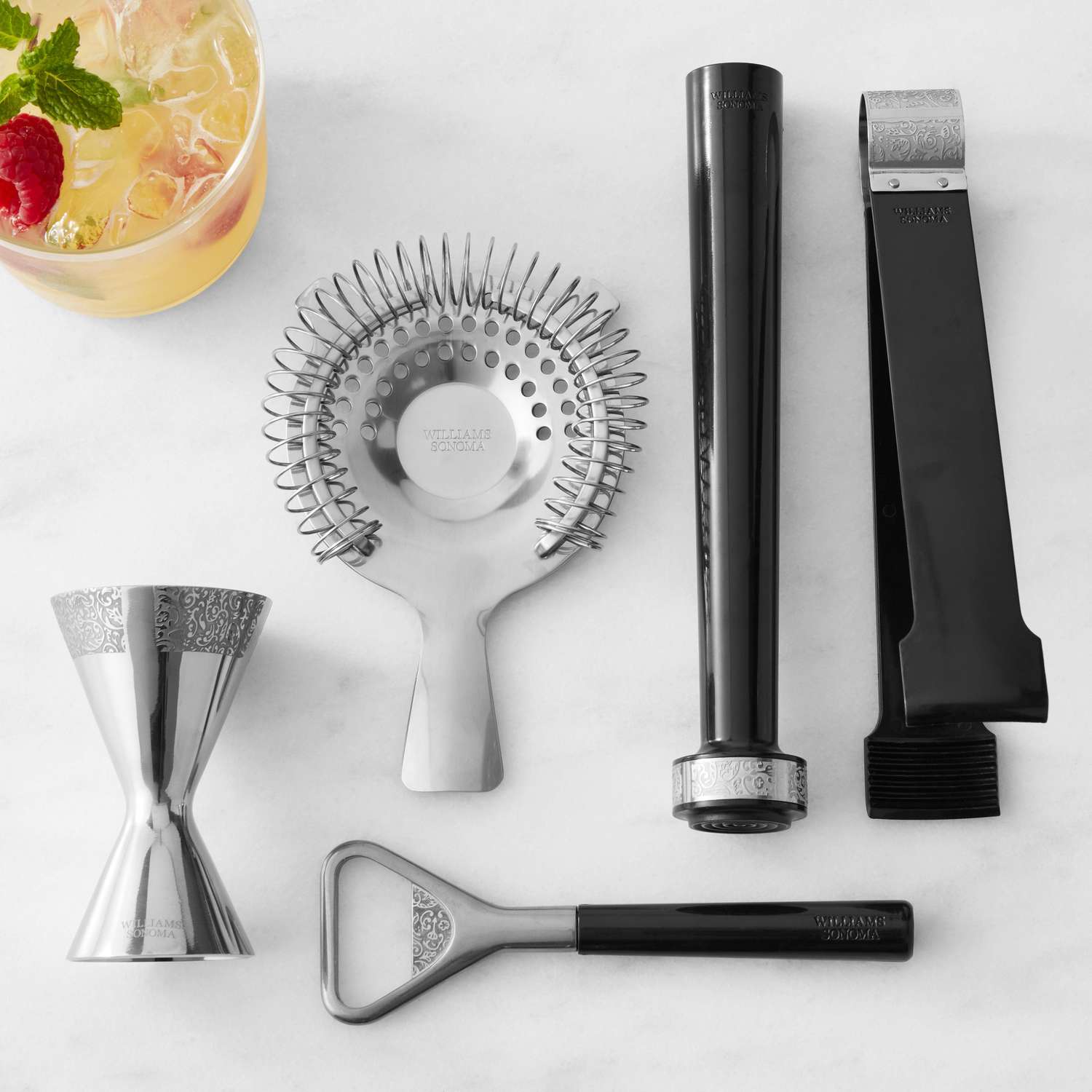 Set of cocktail accessories including a jigger, strainer, muddler, and bottle opener, arranged next to a drink with garnishes