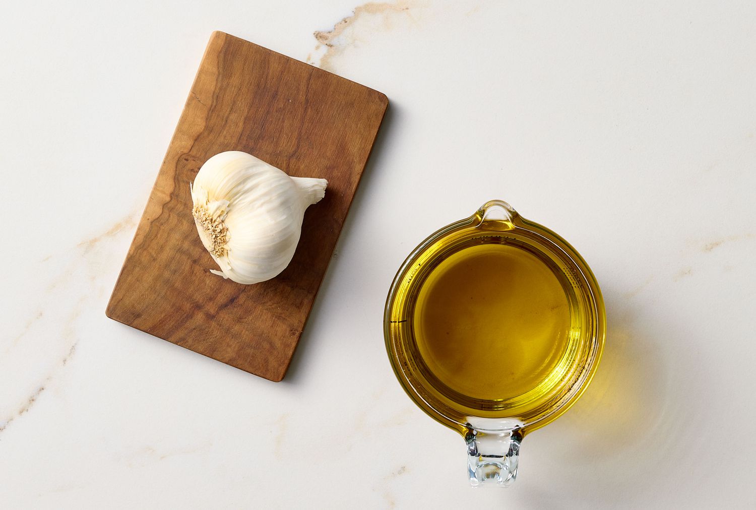 A garlic bulb on a wooden board and a clear bowl of olive oil on a white surface