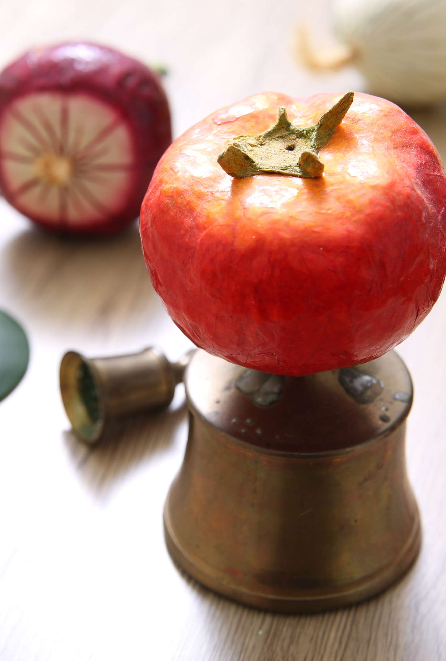 A tomato resting on top of an inverted brass cup with other vegetables in the background