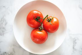 Three ripe red tomatoes on the vine shot in a brightly lit neutral setting.