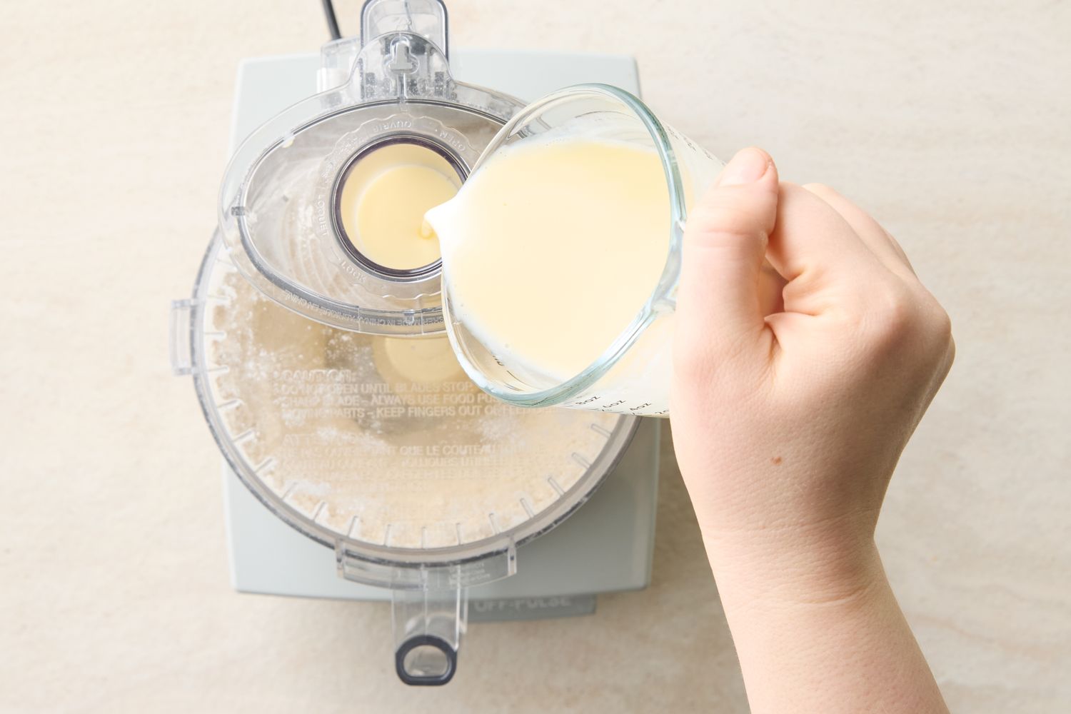 Hand pouring liquid from a measuring cup into a food processor