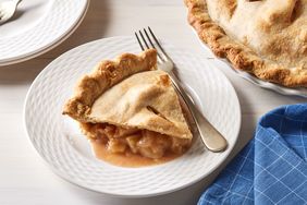 slice of apple pie on white plate with fork, whole pie and blue napkin nearby