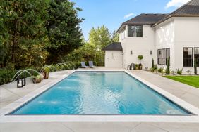 A backyard view featuring a rectangular swimming pool loungers at one end trees and a modern white house on one side