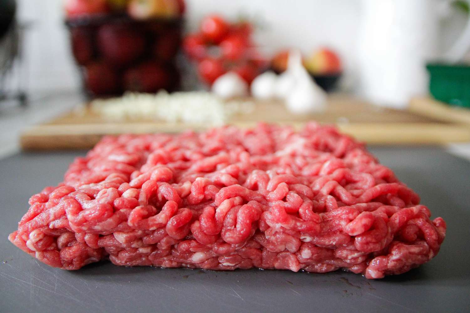 Ground meat portion with blurred kitchen ingredients in the background including cut vegetables and garlic