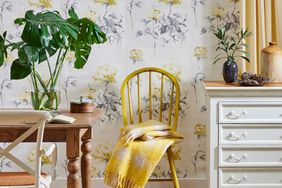 A cozy dining setup with a wooden table a yellow chair draped with a blanket and floral wallpaper in the background a dresser beside