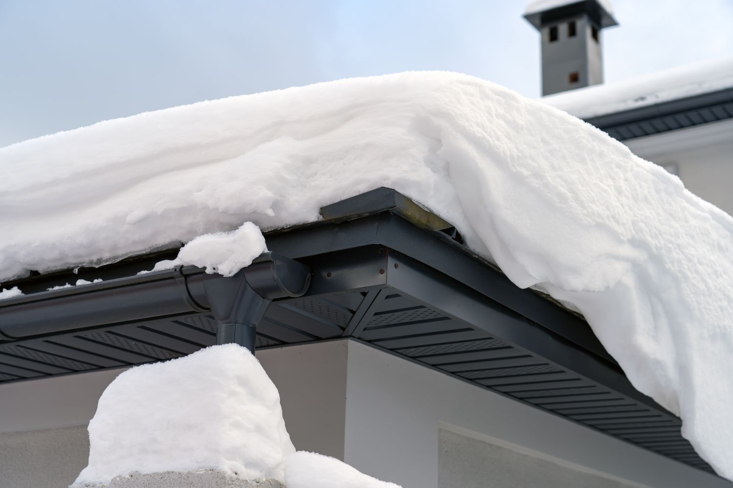 Metal Downpipe system, Guttering System, External downpipes and drainage pipes under snow. Corner of house with roof made of gray metal tiles and gutter covered with thick layer of snow in winter.