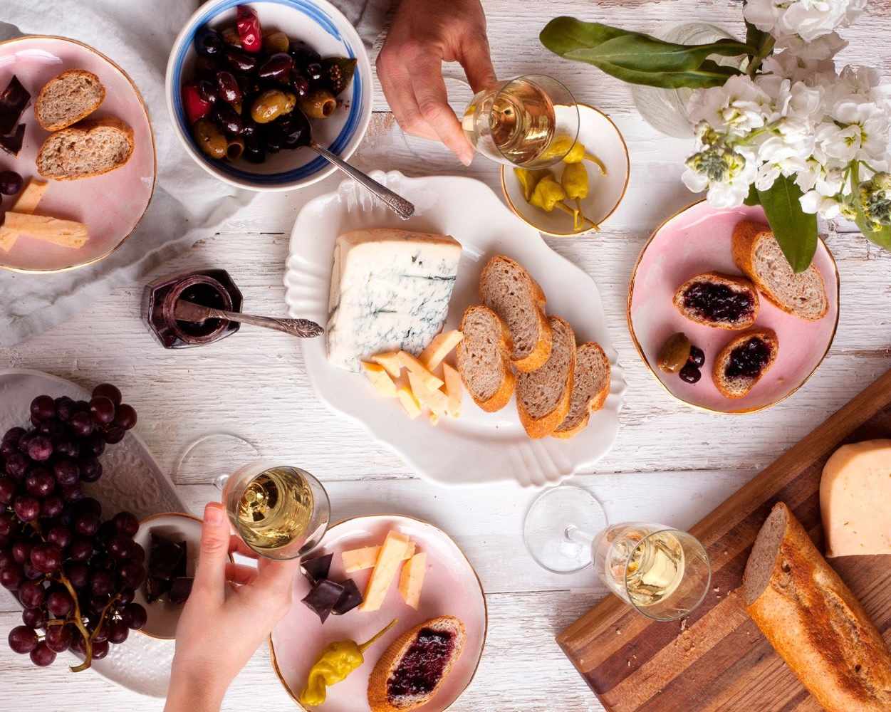 Table set for a dinner party with assorted foods such as bread cheeses and fruits along with glasses of wine
