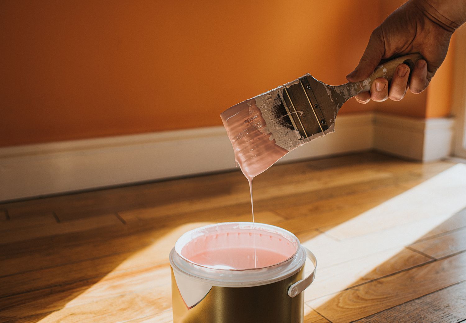 A hand holding a paintbrush with paint dripping into a can in a room with a wooden floor
