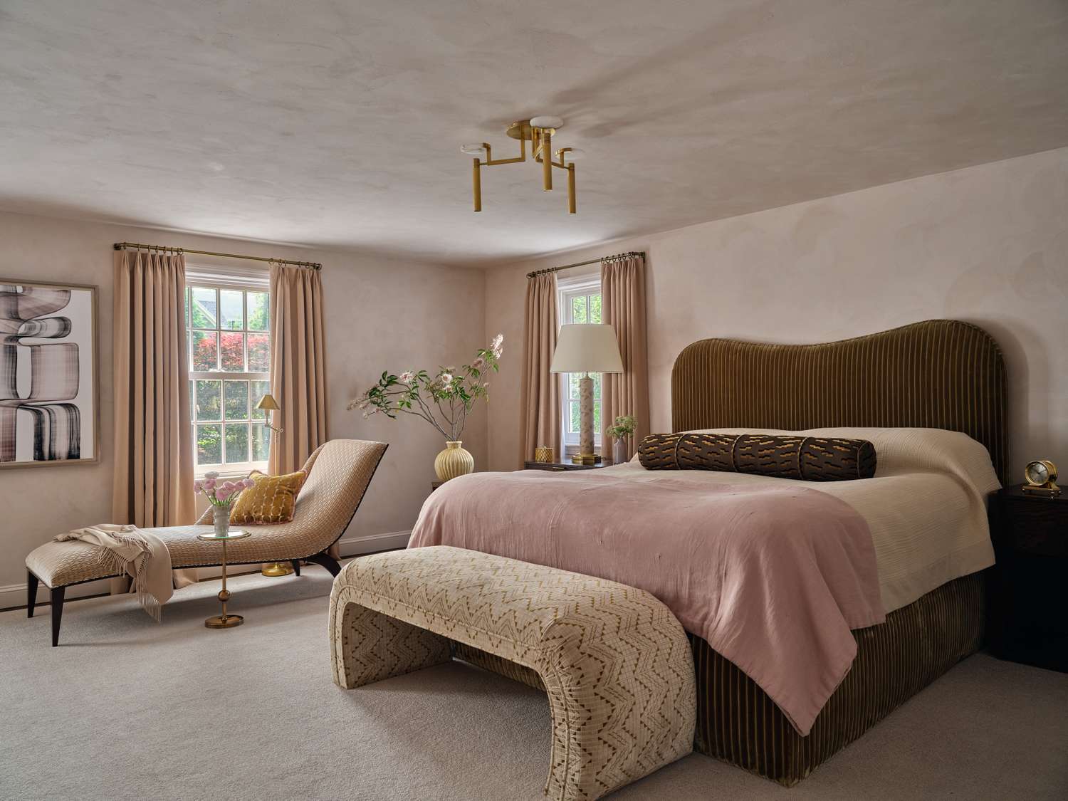 Elegant bedroom with a cushioned bed side table artwork and a chaise lounge near a window with curtains