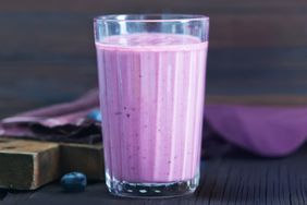 A glass of a purple smoothie with blueberries on the side wooden and cloth surfaces visible
