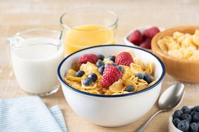 Bowl of cereal topped with raspberries and blueberries surrounded by milk orange juice and bowls of berries and cereal