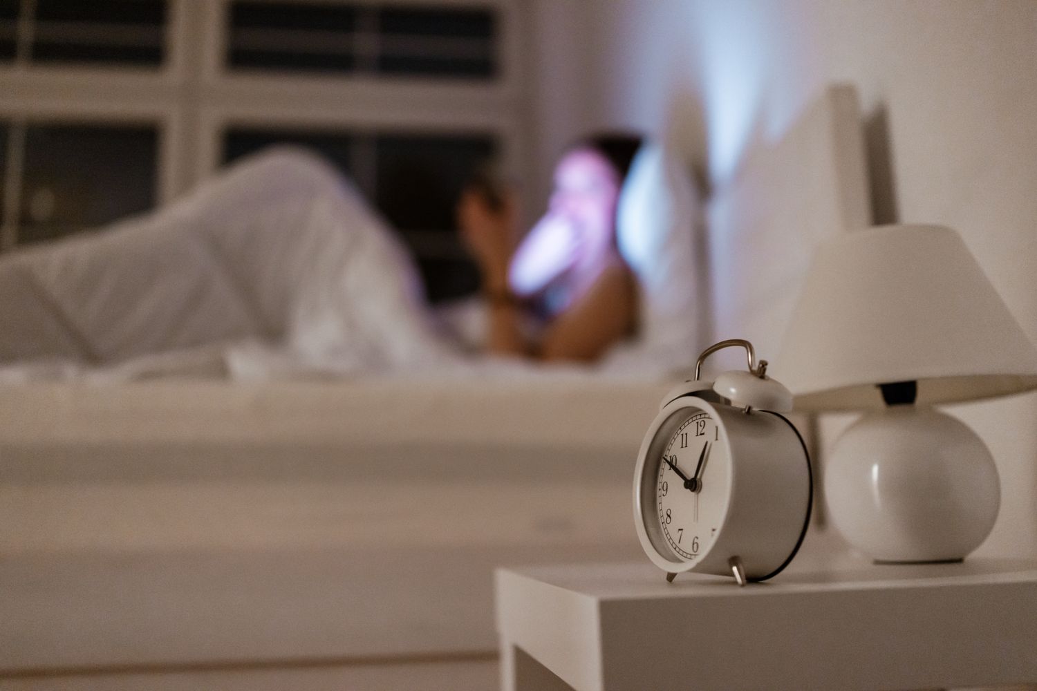 An alarm clock on a bedside table with a blurred figure lying in bed in the background