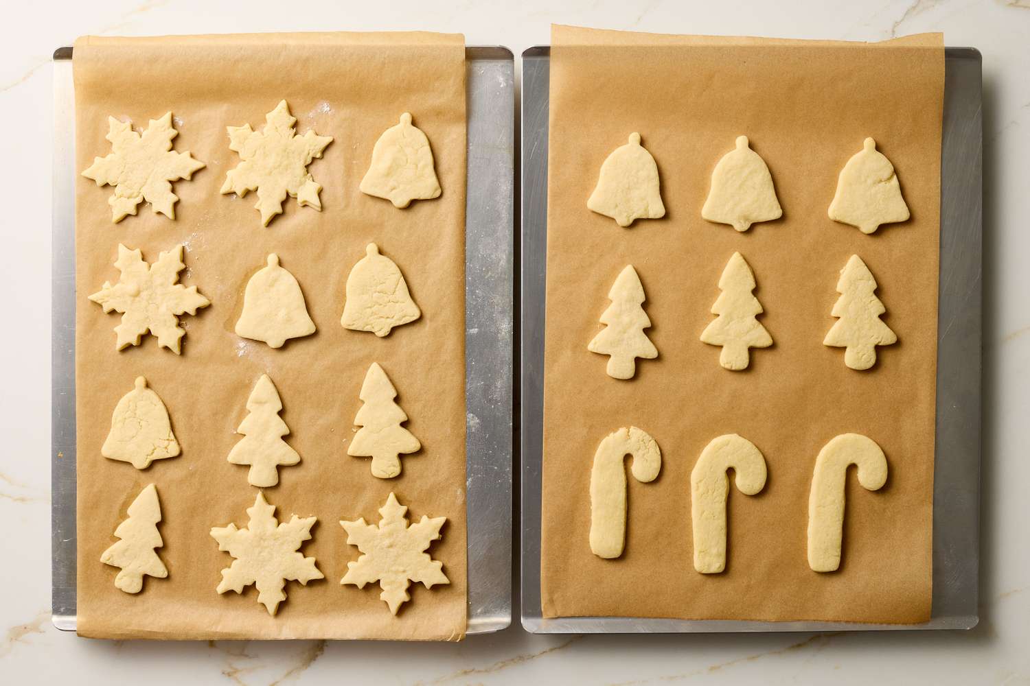 Two trays of unbaked sugar cookies shaped as bells trees snowflakes and candy canes on parchment paper ready for baking