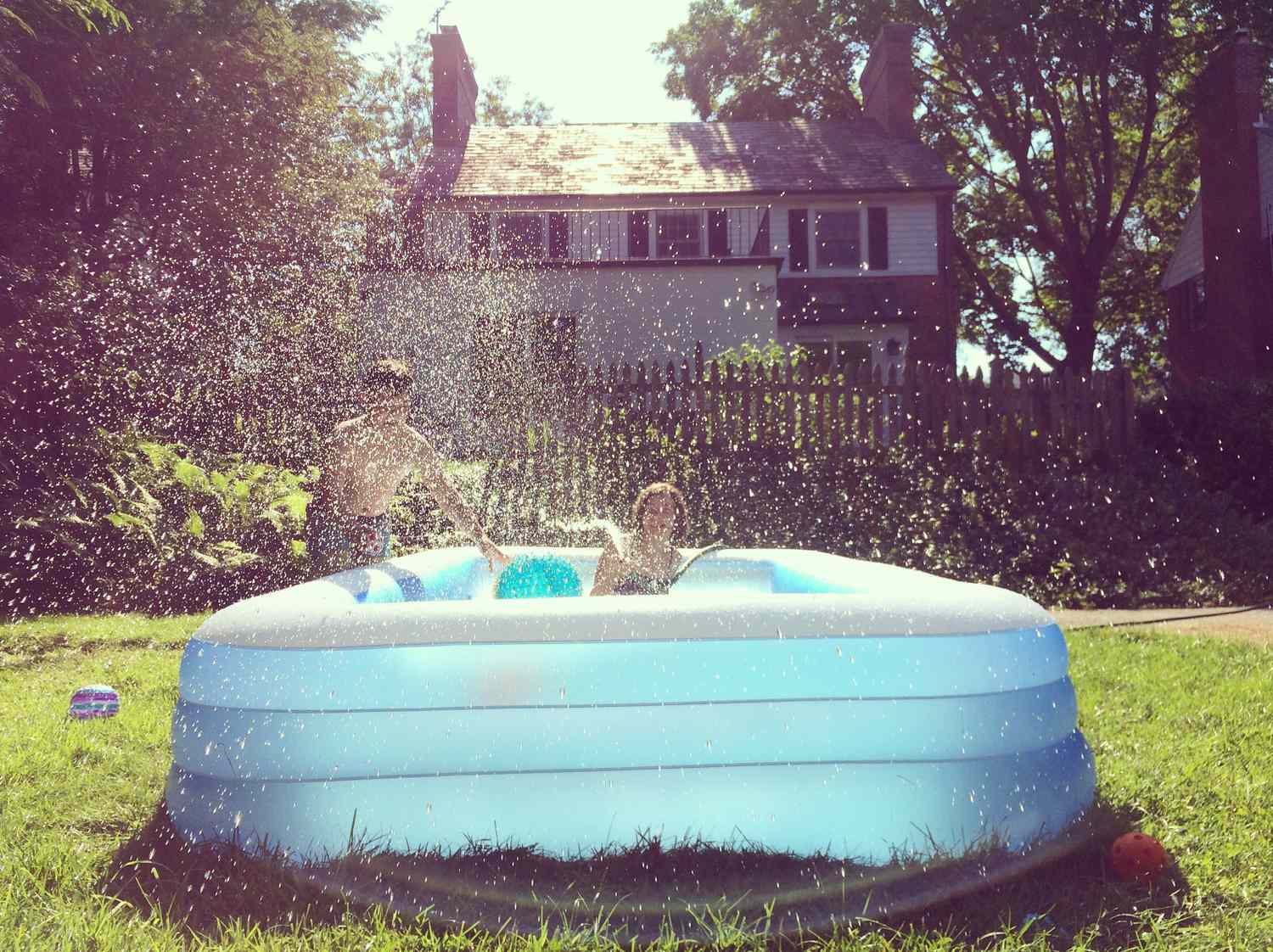 Two children playing in an inflatable pool in a backyard water splashing around them