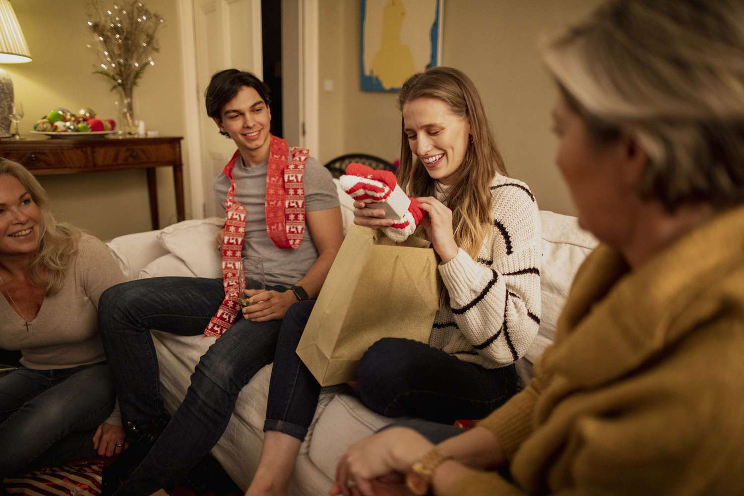 A group of friends are sitting together in the living room at a house party, opening Christmas presents.