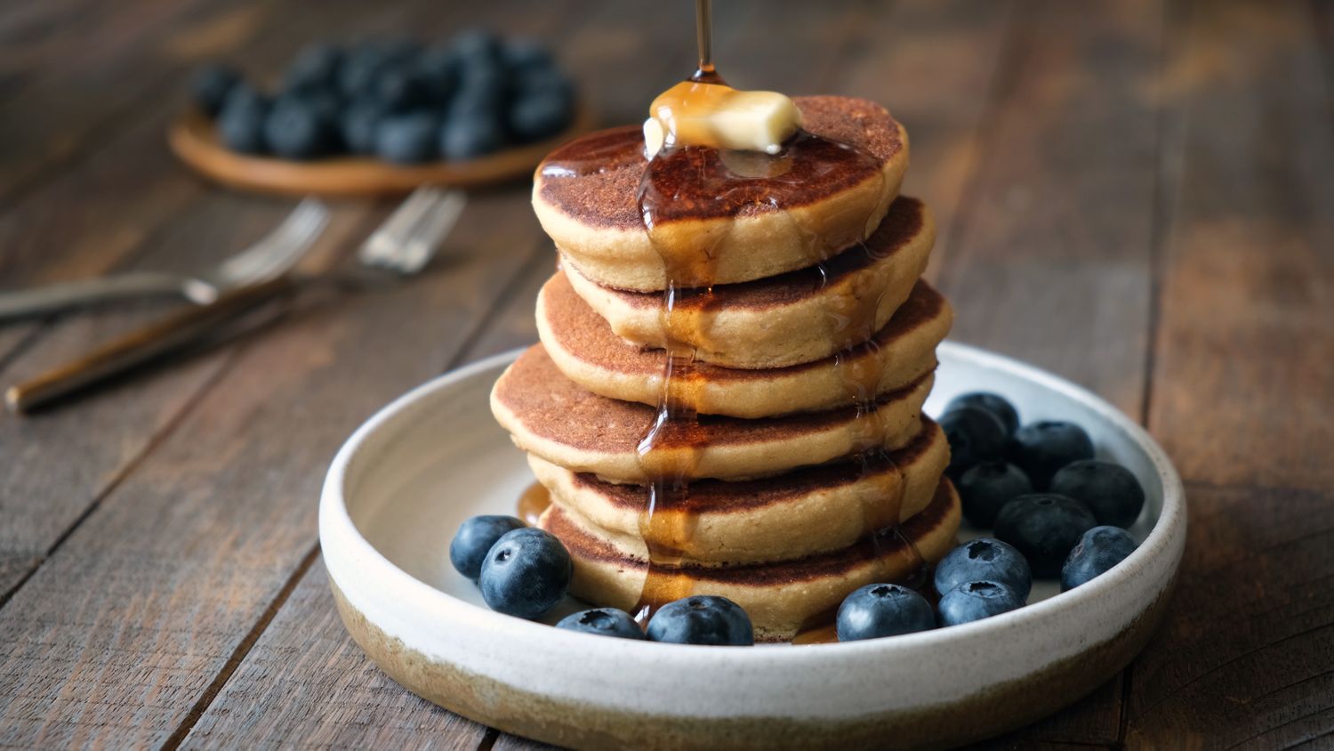 stack of fluffy pancakes on white plate with butter, syrup, and blueberries
