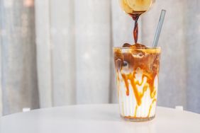 A glass of iced milk with caramel and coffee being poured into it with a straw inserted