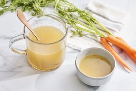 A glass measuring cup and bowl containing chicken stock with fresh carrots and a cloth nearby