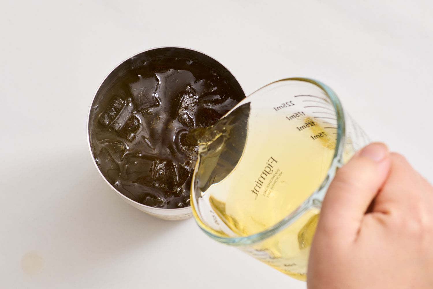 A person pours liquid from a measuring cup into a stainless steel cup filled with ice