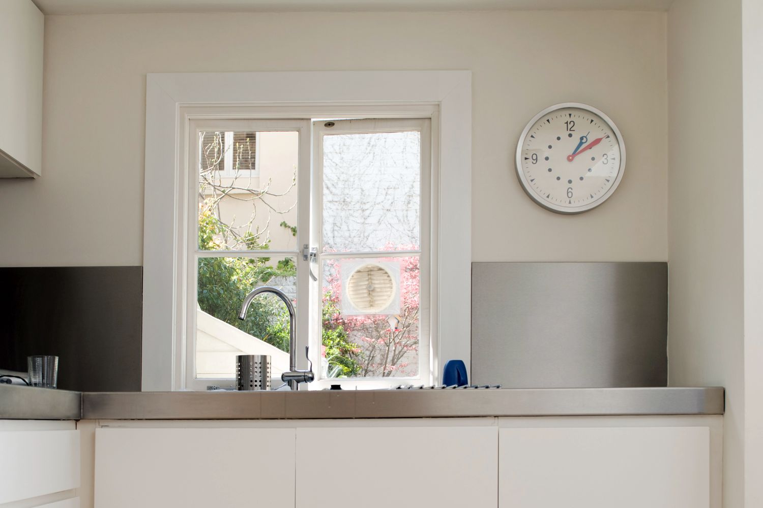 A kitchen sink beneath a window a wall clock is mounted to the right