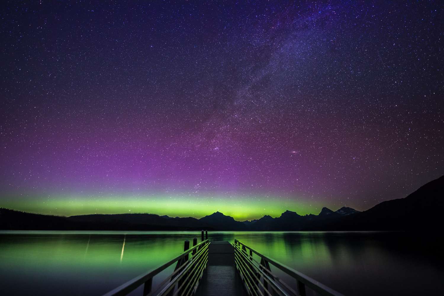 Northern lights and starry sky viewed over a lake with a pier extending from the foreground to the water's edge