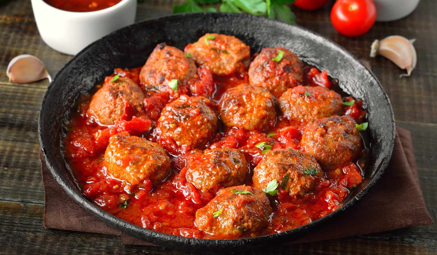A pan of meatballs cooked in red sauce garnished with parsley and surrounded by ingredients like garlic and tomatoes