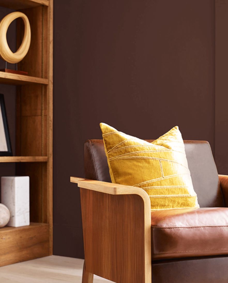 A wooden armchair with a leather cushion and a yellow pillow in a room with dark walls and a wooden shelf