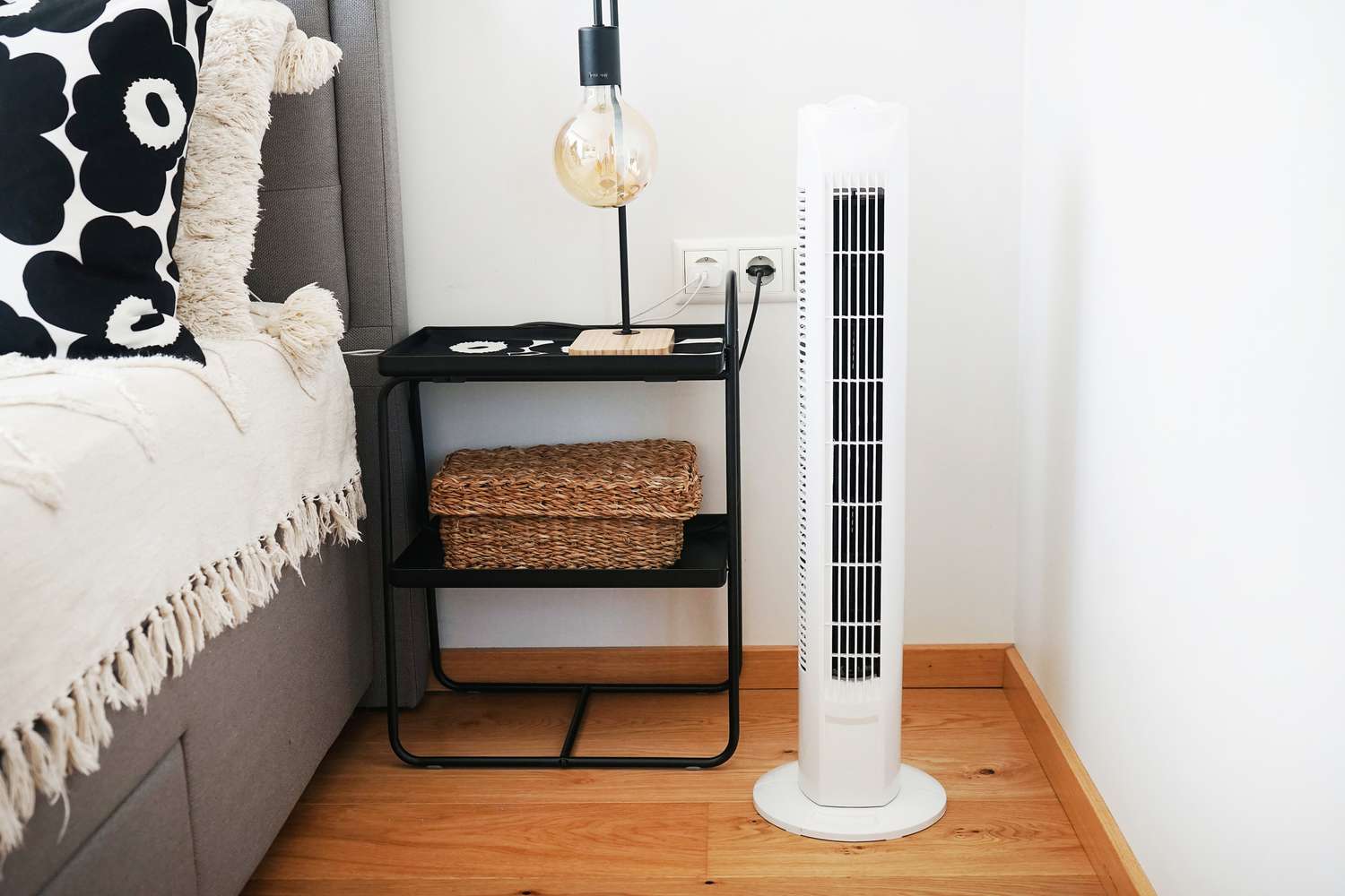 A bedside table with a lamp, baskets, and a tower fan nearby on a wooden floor