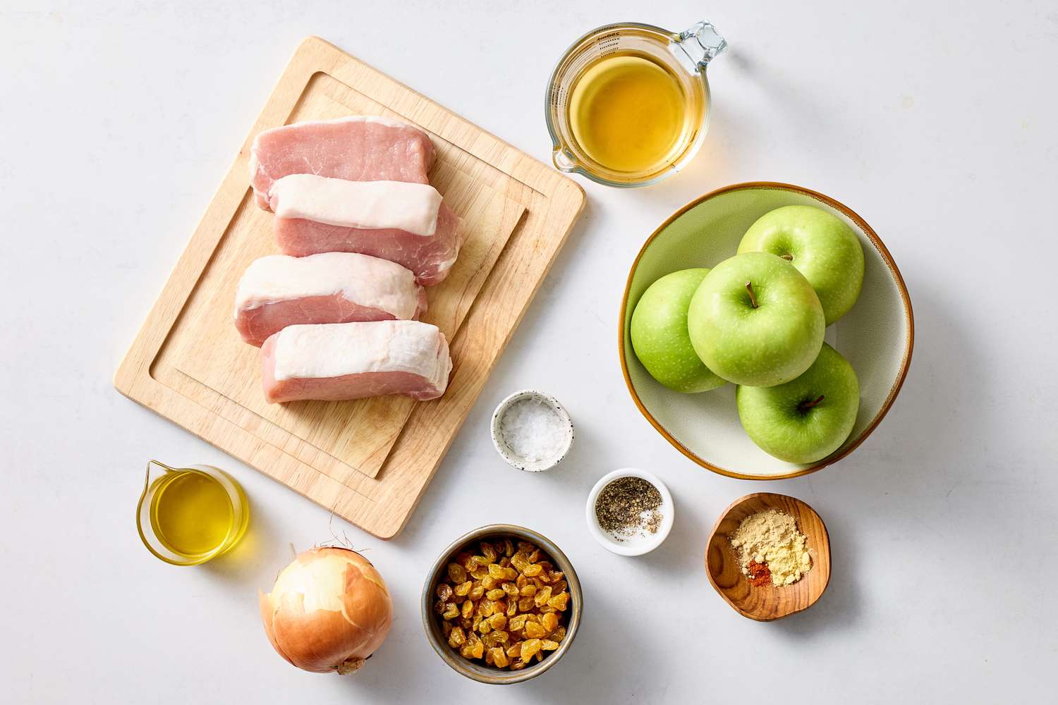 Ingredients for a dish including boneless pork chops apples onion and seasonings arranged on a surface