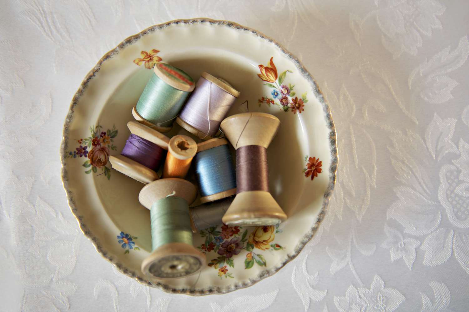 A porcelain bowl containing various spools of thread on a patterned tablecloth