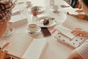 People sitting around a table reading books cups and plates are on the table