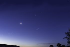 Night sky with celestial bodies and a crescent moon visible above a silhouette of treetops and hills