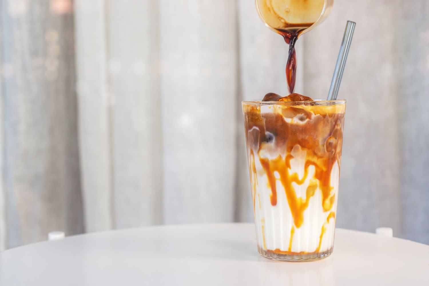 A glass of iced milk with caramel and coffee being poured into it with a straw inserted
