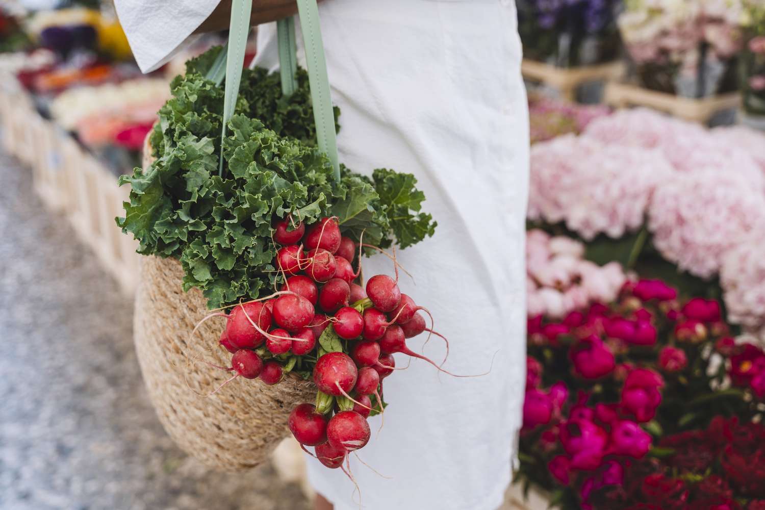 Close-up of a bag filled with fresh kale and radishes, held at an outdoor farmers market with flowers in the background