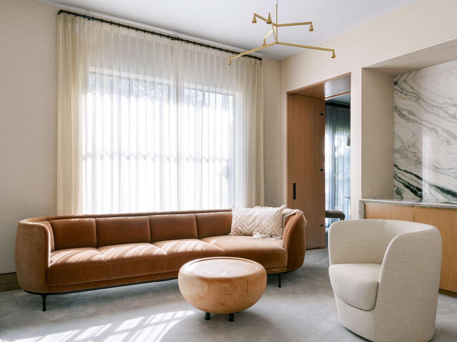 Minimalist living room by Lambrakos Studio: taupe sectional sofa, beige curved chair, wooden ottoman, and brass chandelier. Marble wall, sheer curtains on large windows, light wood floor, and natural light create a calm, stylish space.