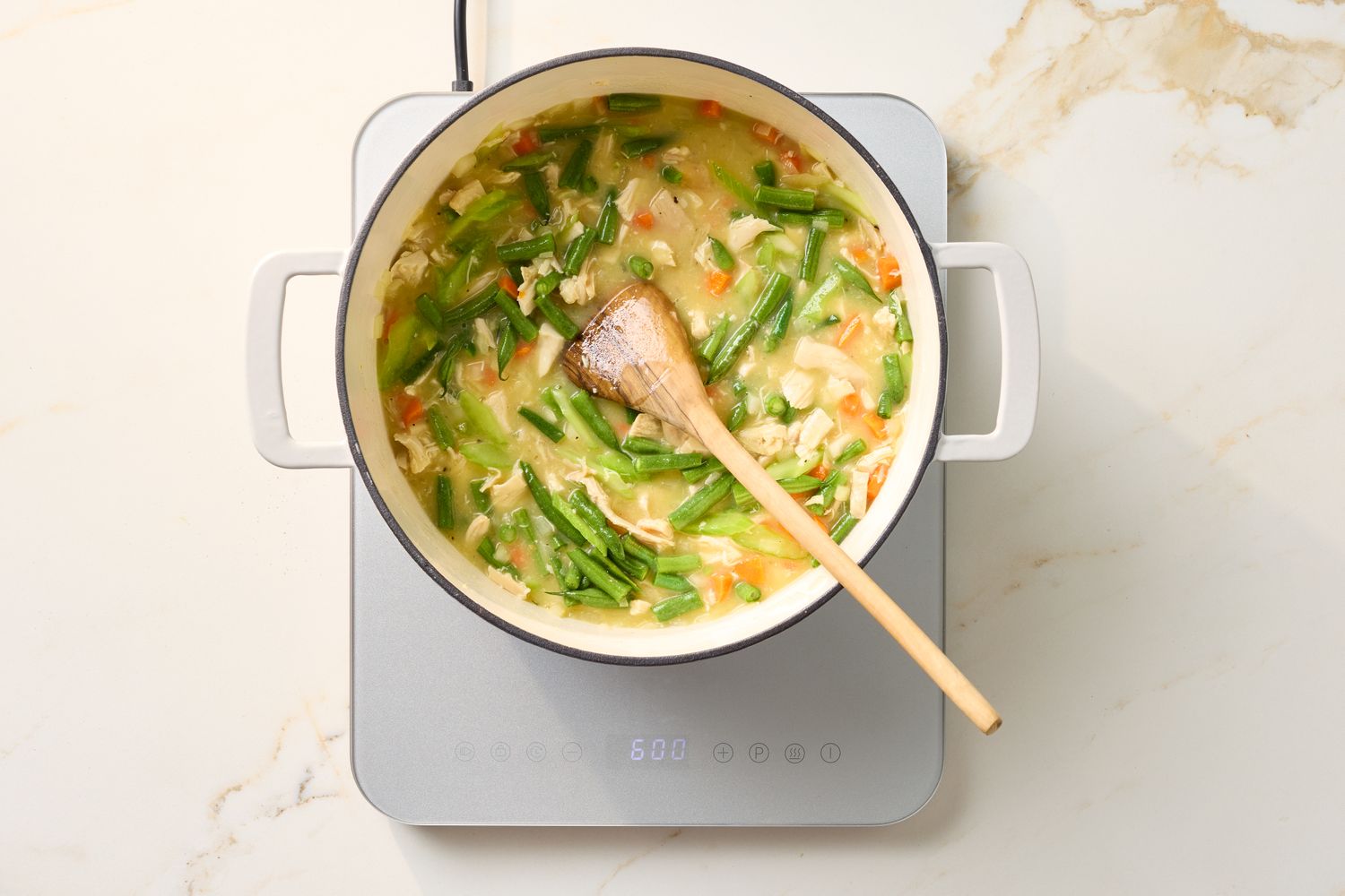 A pot on a stovetop containing chicken and dumplings with vegetables being stirred with a wooden spoon