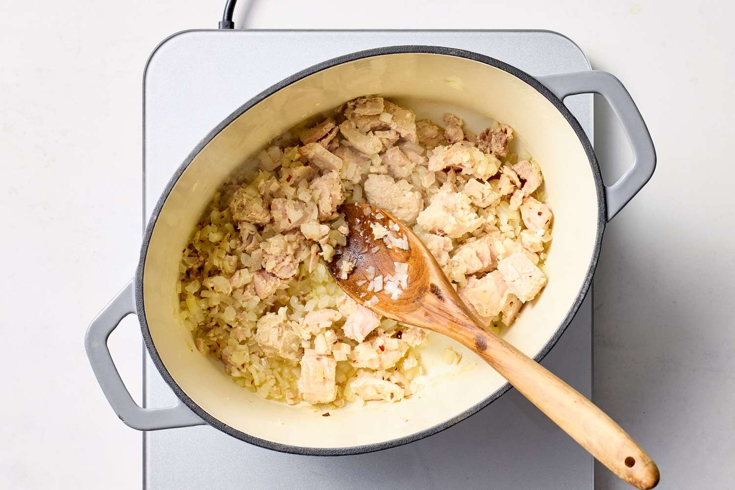 A pot with sauting onions and chunks of tuna stirred with a wooden spoon