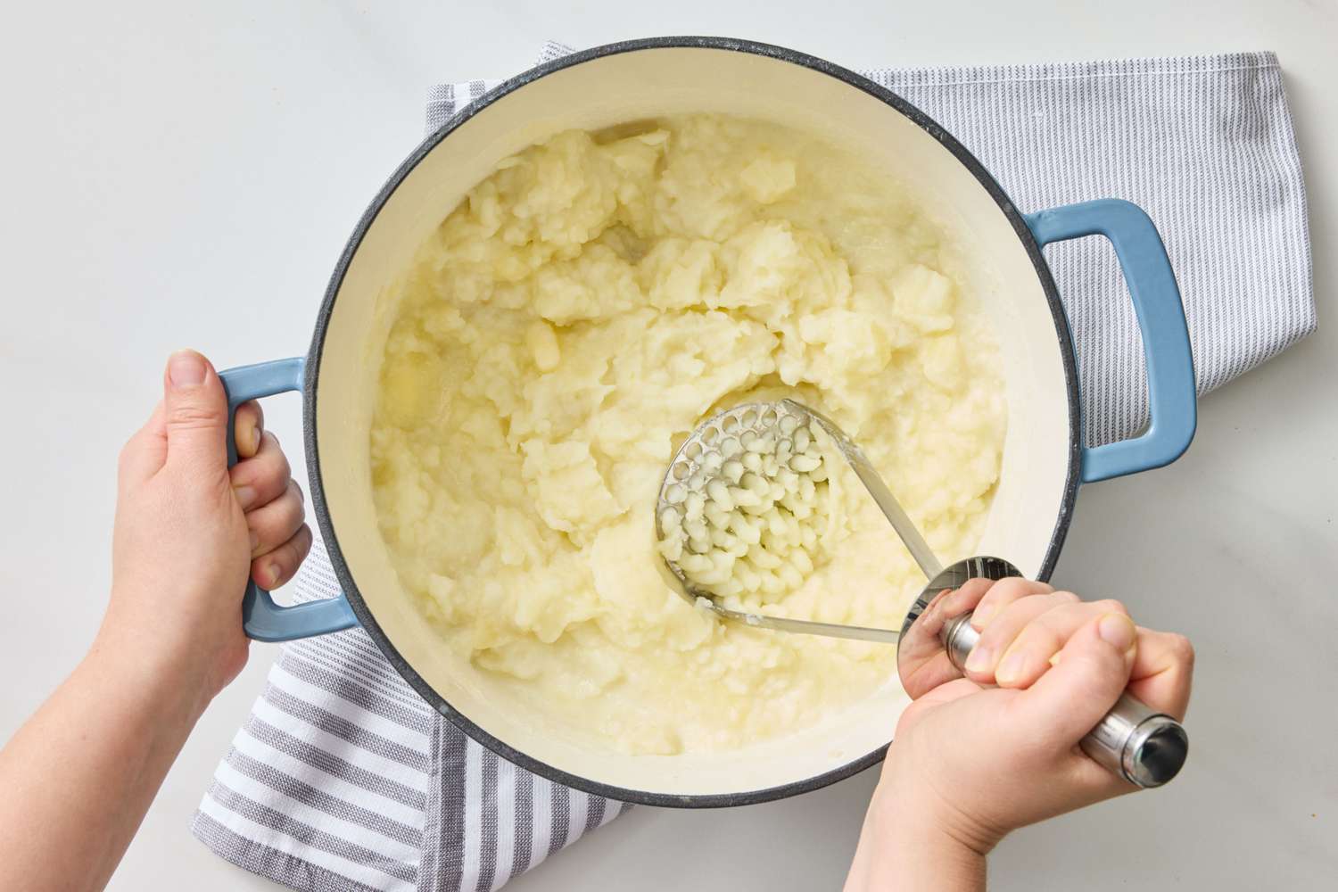 Hands mashing potatoes in a pot with a metal masher