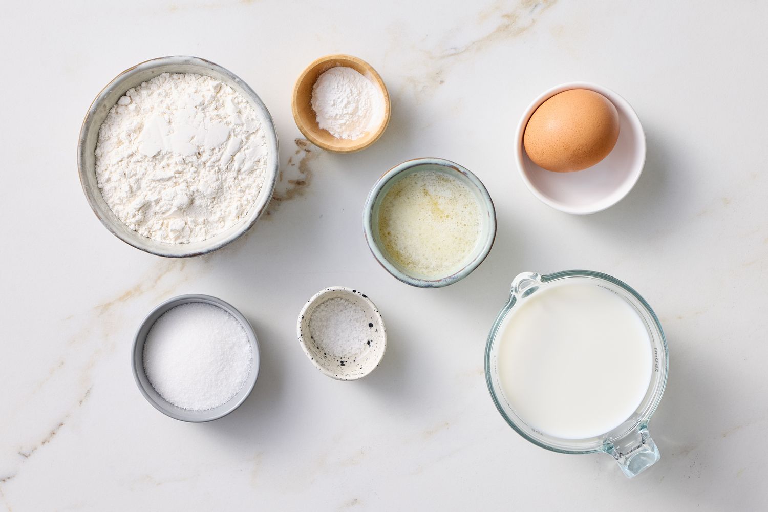 Ingredients for pancakes on a white surface including flour sugar salt baking powder milk a whole egg and melted butter